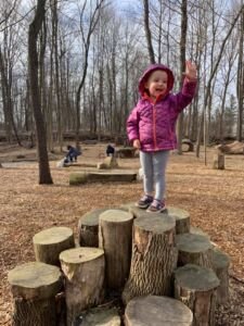 Toddler girl in pink coat standing on stump of wood and waving hand like a queen