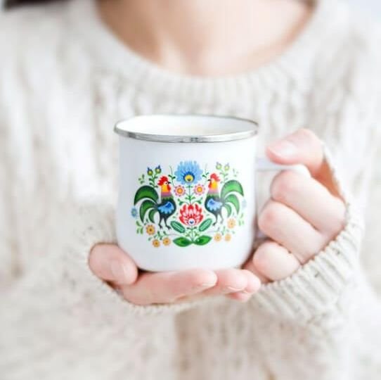 Woman in white sweater holding a white mug painted with flowers and roosters.