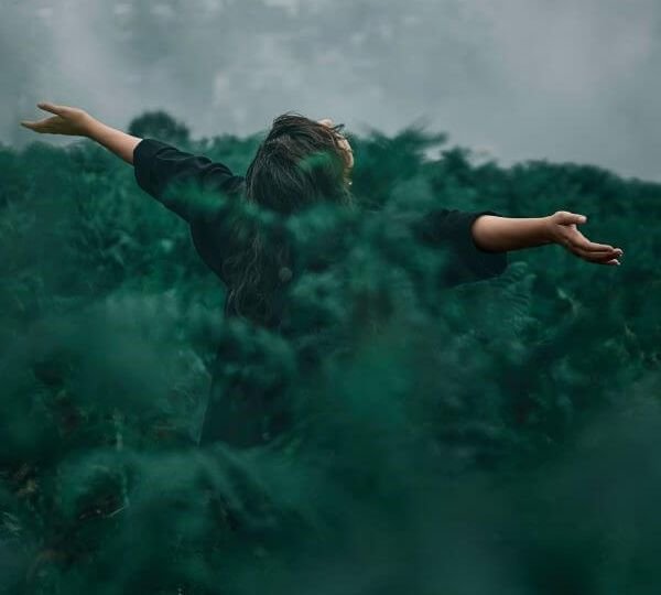 A woman in a black shirt stands amid ferns. Her arms are stretched out to the right and left, and her head is tilted upwards towards the sky.