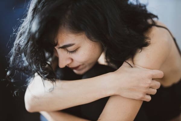 Woman with black hair sits on ground hugging her knees to her chest. She looks sad.