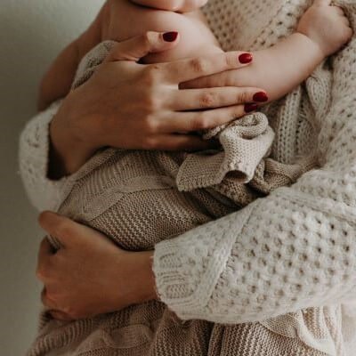 Close-up shot of a woman with red fingernail polish holding an infant in her arms. She is wearing a white sweater and the baby is wrapped in a beige blanket.